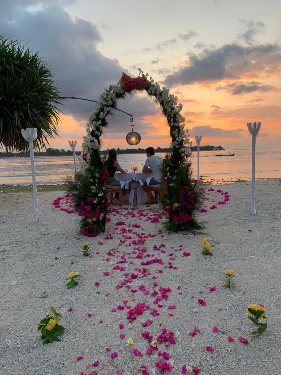 Un couple est assis à une table placée sous une arche fleurie, sur une plage, avec une vue sur le coucher de soleil.