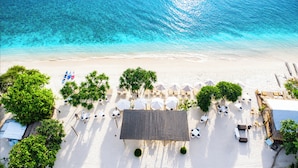 Vue aérienne d’un complexe hôtelier en bord de mer, avec son sable blanc, ses eaux turquoise et limpides, et une rangée de parasols blancs et de chaises longues.