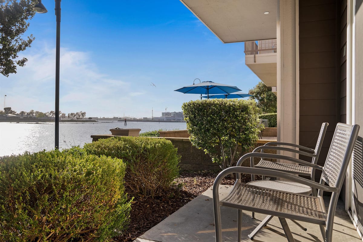 A patio with metal chairs and a blue umbrella, overlooking a body of water with a view of buildings and a bridge in the distance.