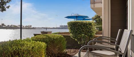 A patio with metal chairs and a blue umbrella, overlooking a body of water with a view of buildings and a bridge in the distance.