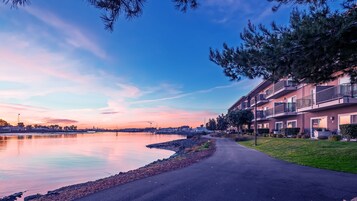 Una zona residencial frente al agua, con un paseo pavimentado a lo largo de la costa, edificios modernos de varios pisos con balcones y un cielo despejado.