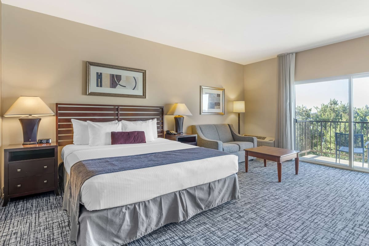 A hotel room with a large bed, two bedside tables with lamps, a chair, a small table, and a balcony with a view.