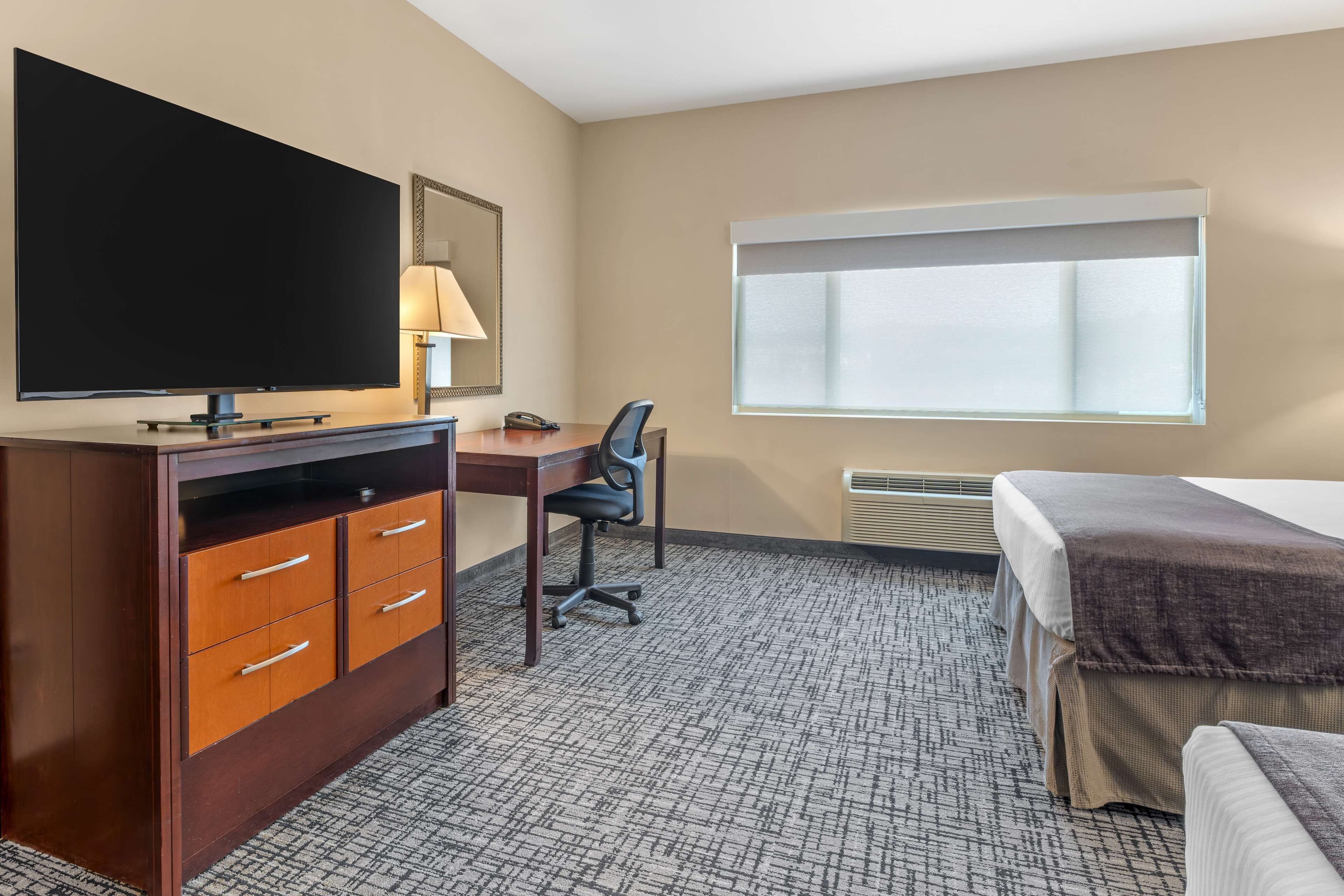 Une chambre d’hôtel dotée d’une grande télévision à écran plat, d’un bureau avec une chaise, d’un lit avec une couverture et d’une fenêtre avec des stores.