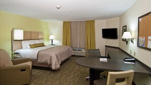 A hotel room with a bed, two chairs, a desk, a television, and a bulletin board.