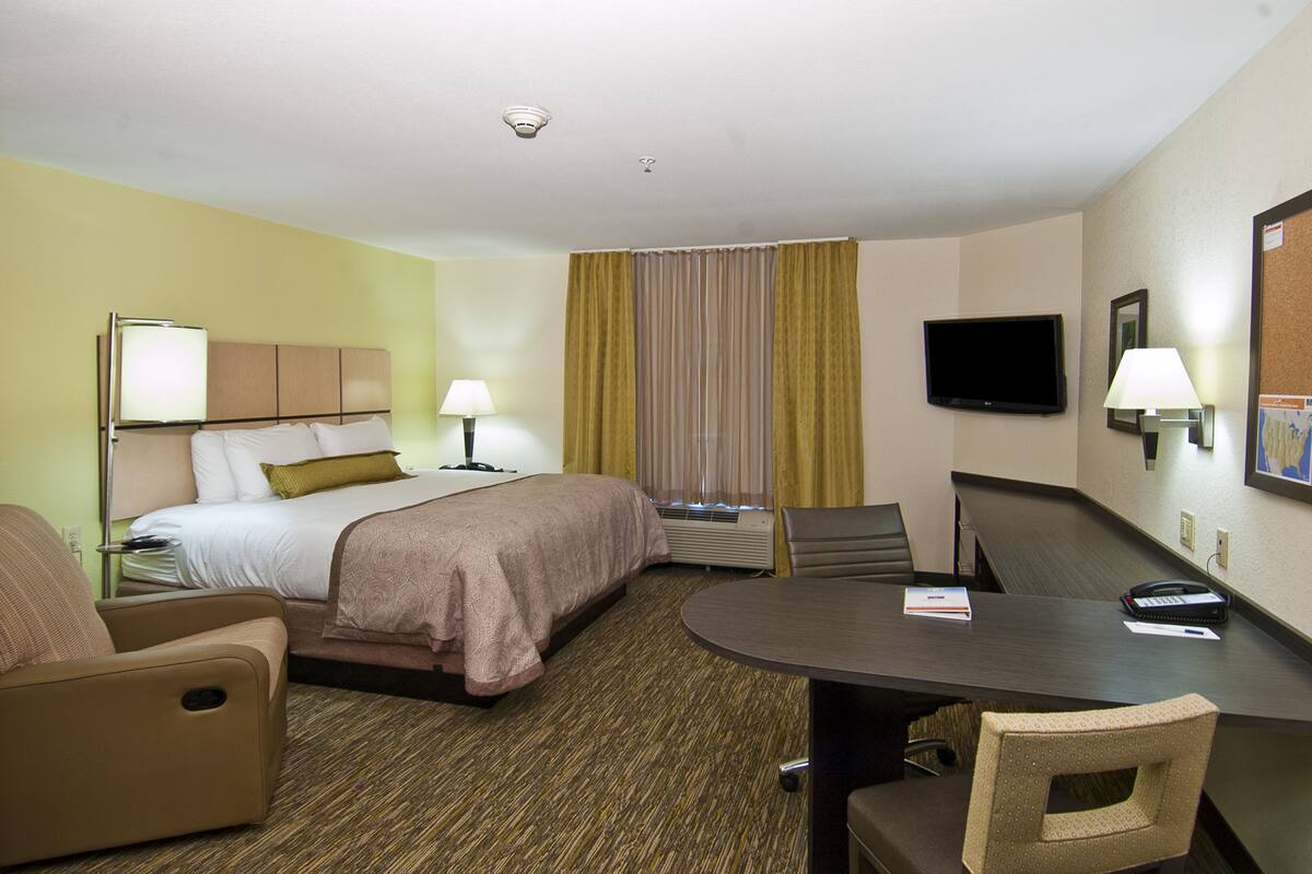 A hotel room with a bed, two chairs, a desk, a television, and a bulletin board.