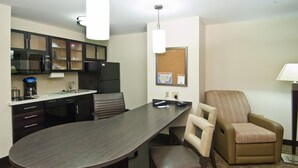 A hotel room with a dark wood dining table, a brown leather chair, a microwave, a refrigerator, and a corkboard on the wall.
