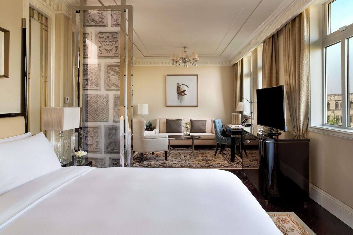 A hotel room with a large bed, a desk with a computer, a chair, a sofa, and a TV.