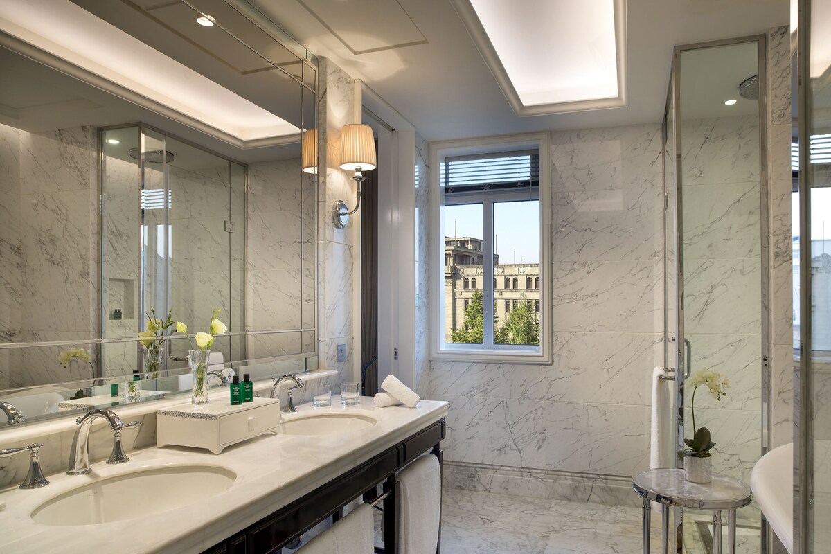 Ein modernes Badezimmer mit zwei Waschbecken, einem großen Spiegel und einem Fenster mit Blick auf Gebäude.