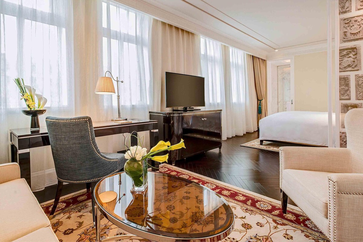 Habitación de hotel con cama, escritorio con televisor, silla, mesa de centro y ventana grande con cortinas.