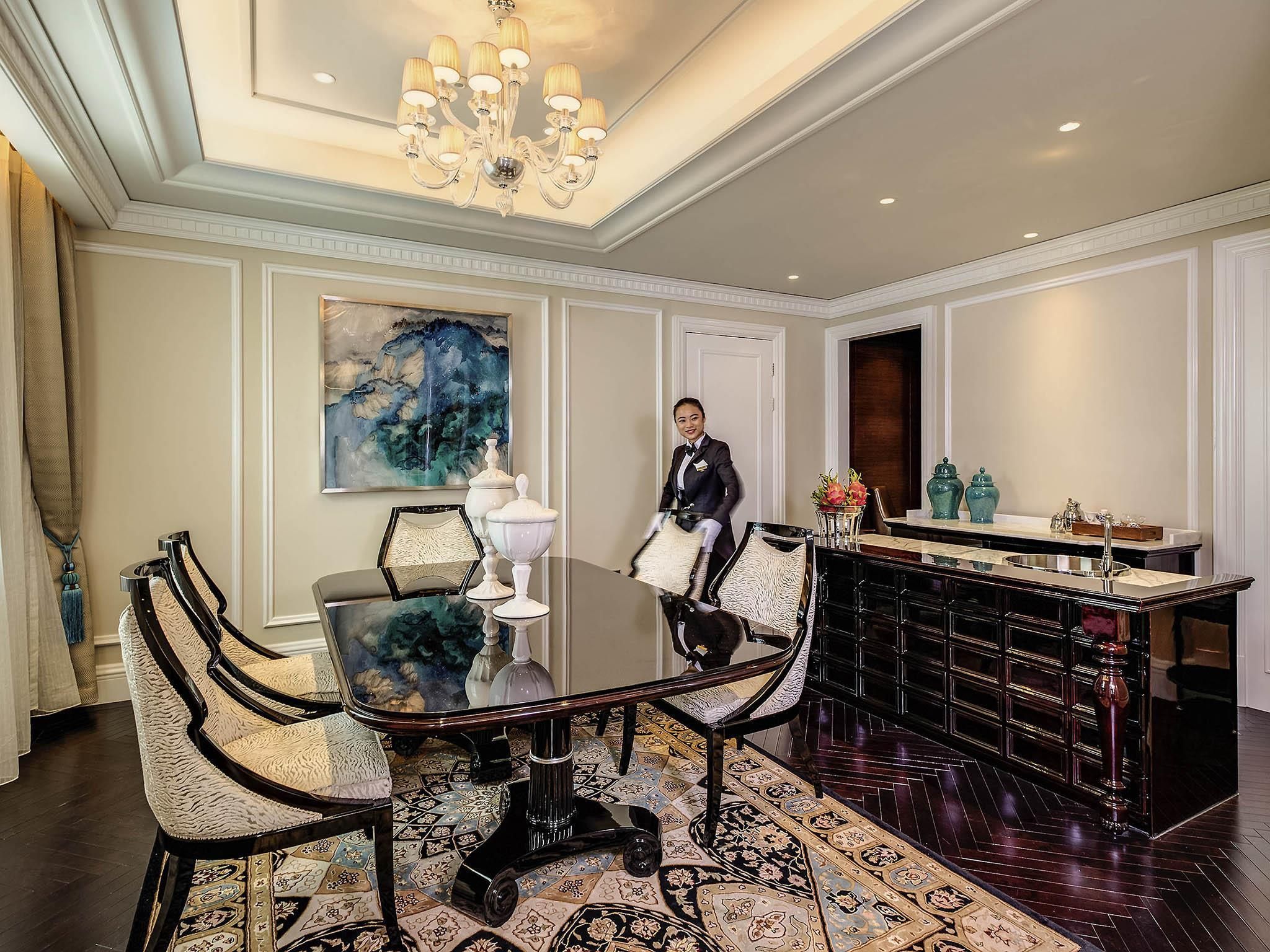 A hotel room with a dining table, chairs, a chandelier, a painting, and a bar area.