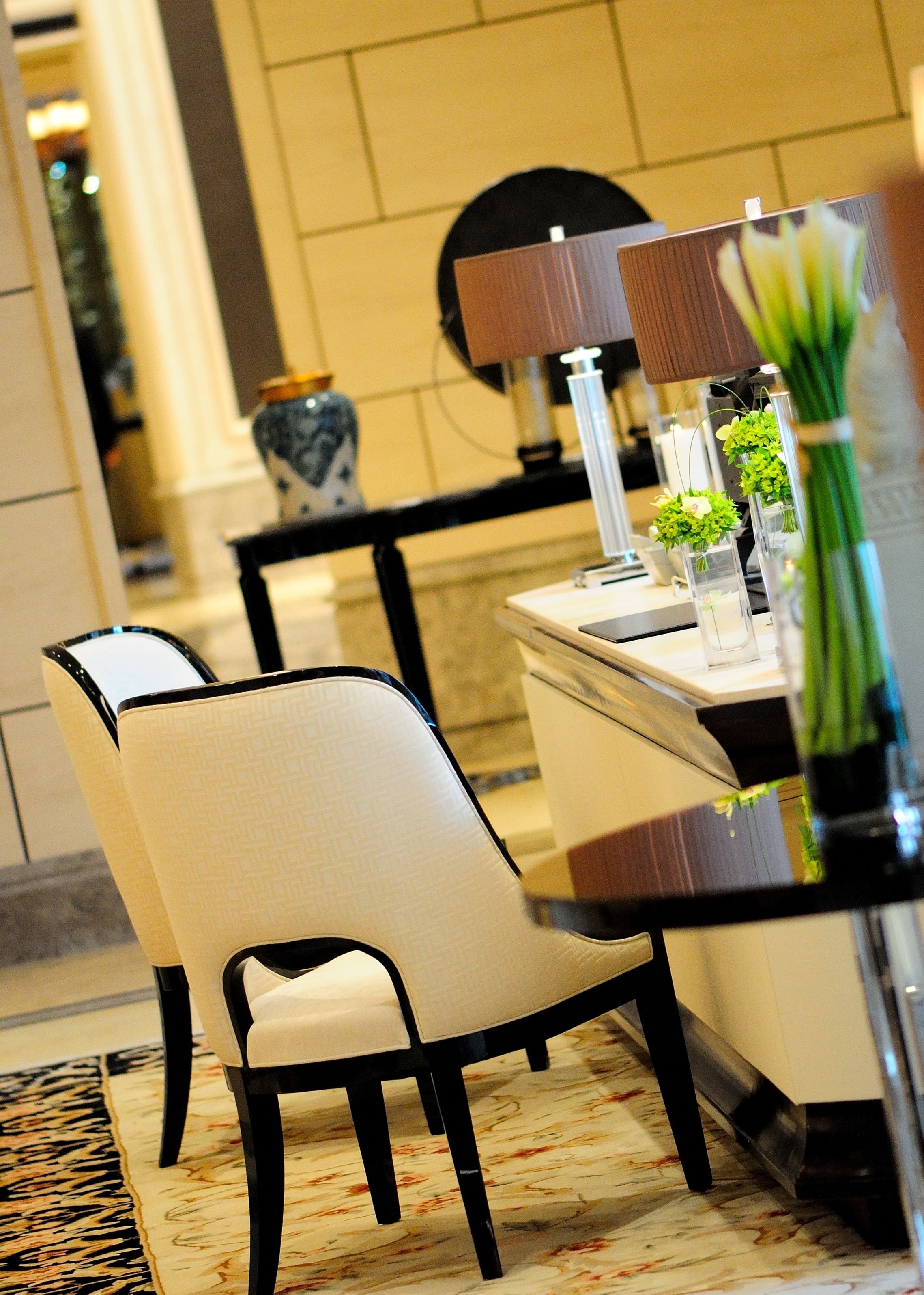 A dining area with a table set for two, featuring a vase with green flowers, a lamp, and a decorative plate.