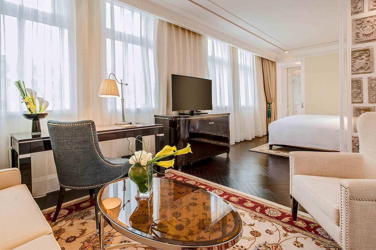 Habitación de hotel con cama, escritorio con televisor, silla, mesa de centro y ventana grande con cortinas.