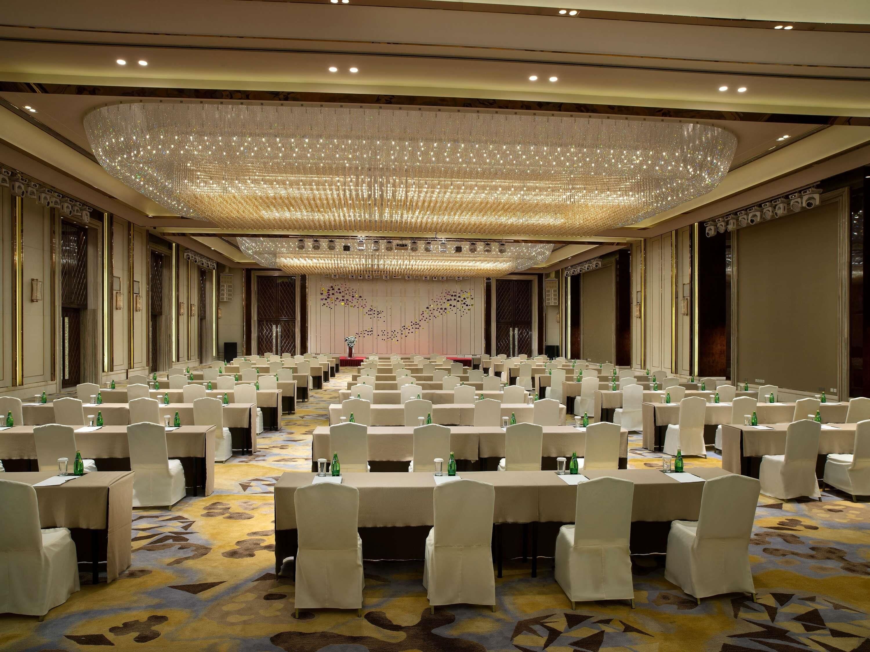 A large conference room with rows of tables and chairs, a decorative ceiling, and a stage with a floral arrangement.