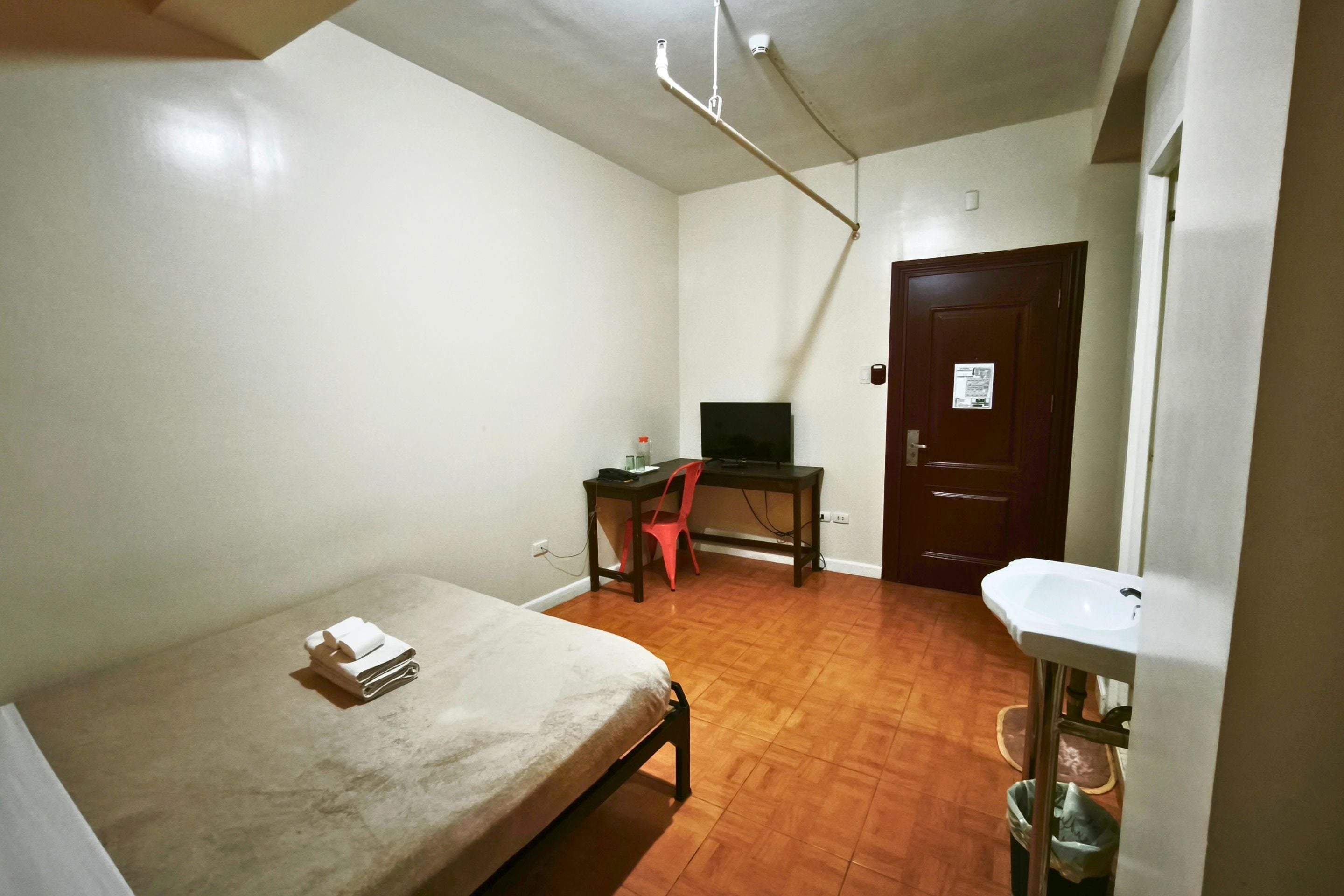 A small, sparsely furnished room with a single bed, a desk with a chair, a television, and a door with a notice.