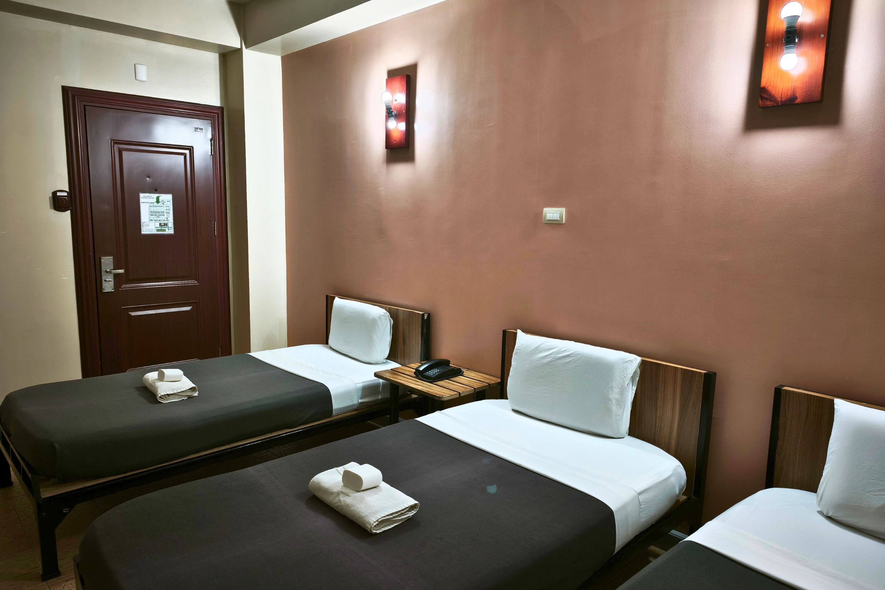 A hotel room with two single beds, a wooden door, a bedside table with a phone, and a wall-mounted light fixture.