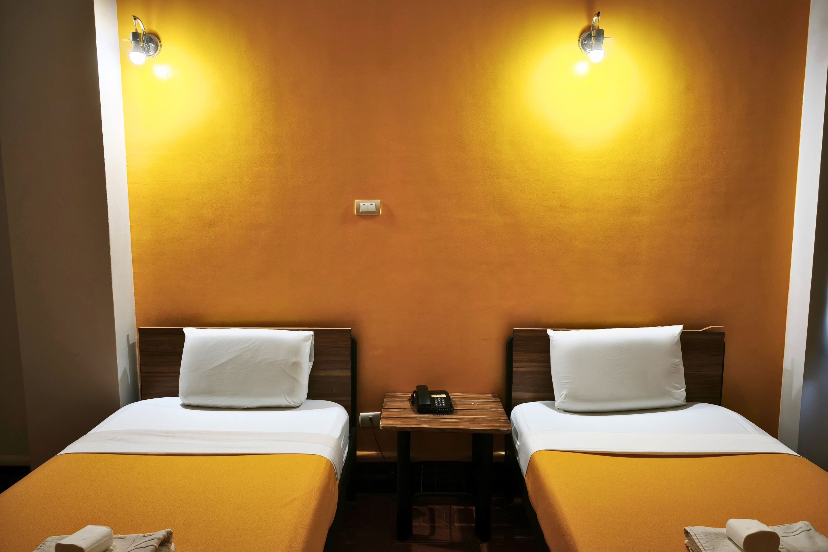 A hotel room with two single beds, a small table with a telephone, and two wall-mounted light fixtures.