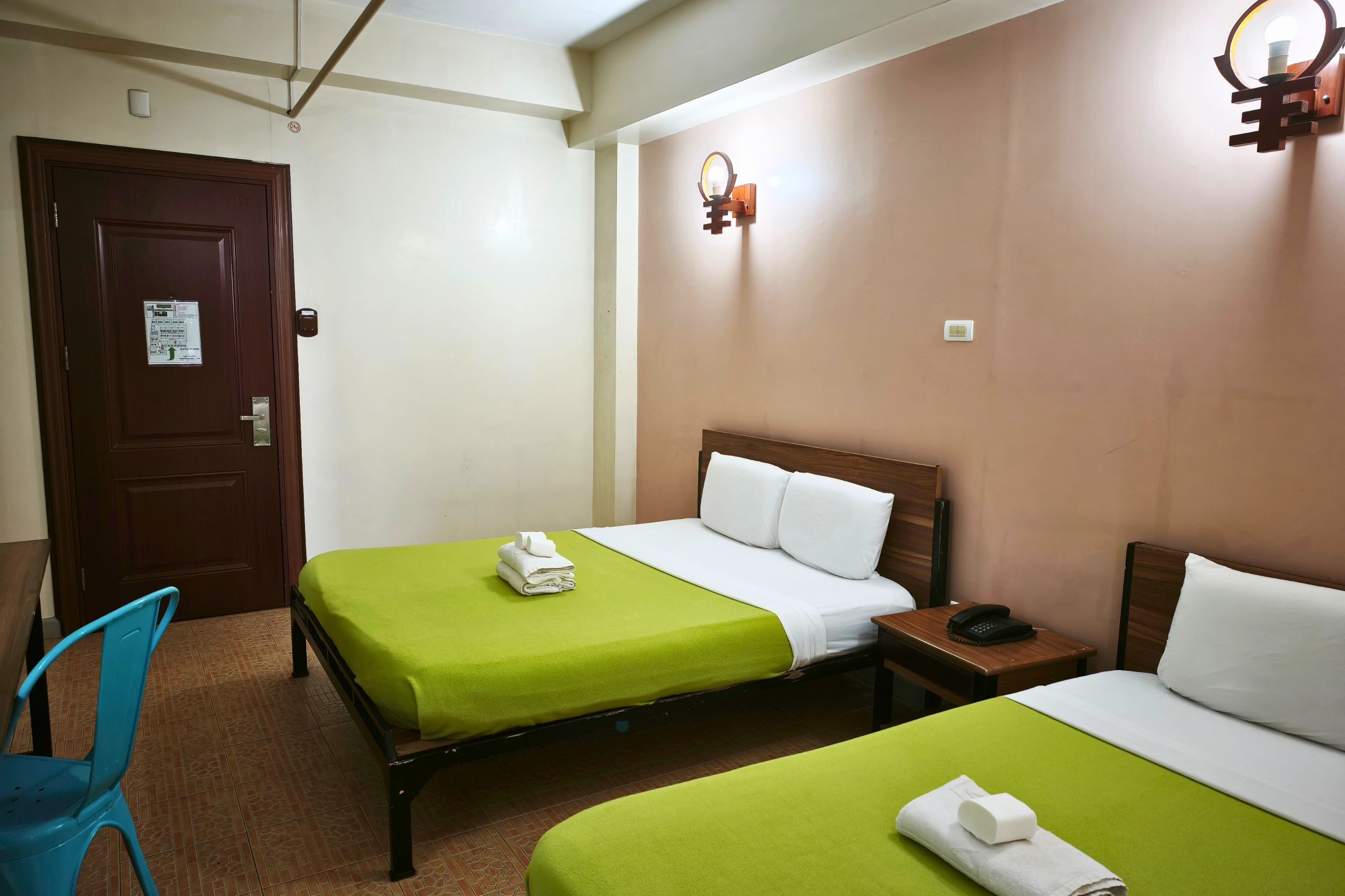 Habitación de hotel con dos camas, una puerta de madera, una mesita con teléfono, y una lámpara fijada a la pared.