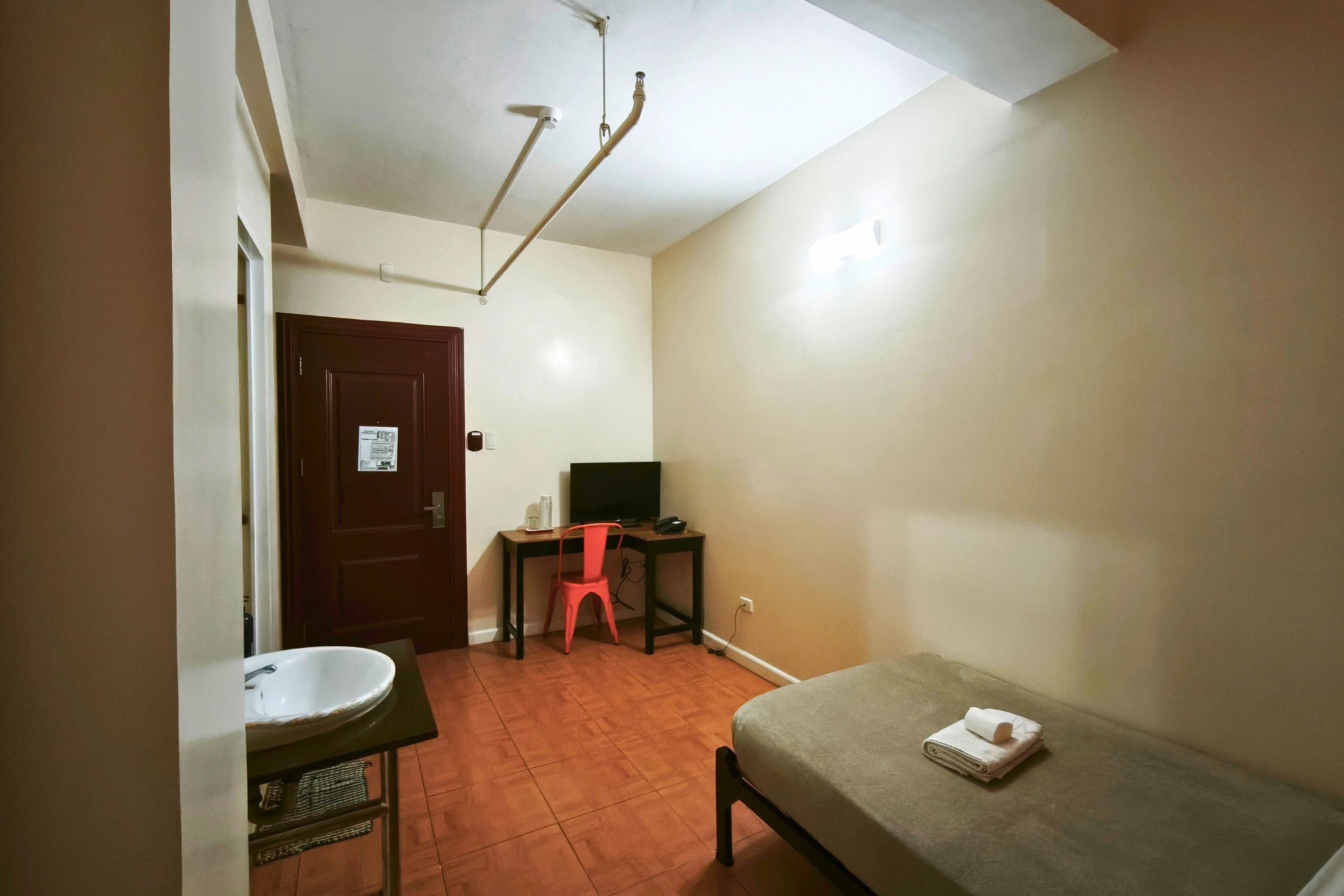 A small room with a bed, a desk with a chair, a television, and a bathroom with a sink.