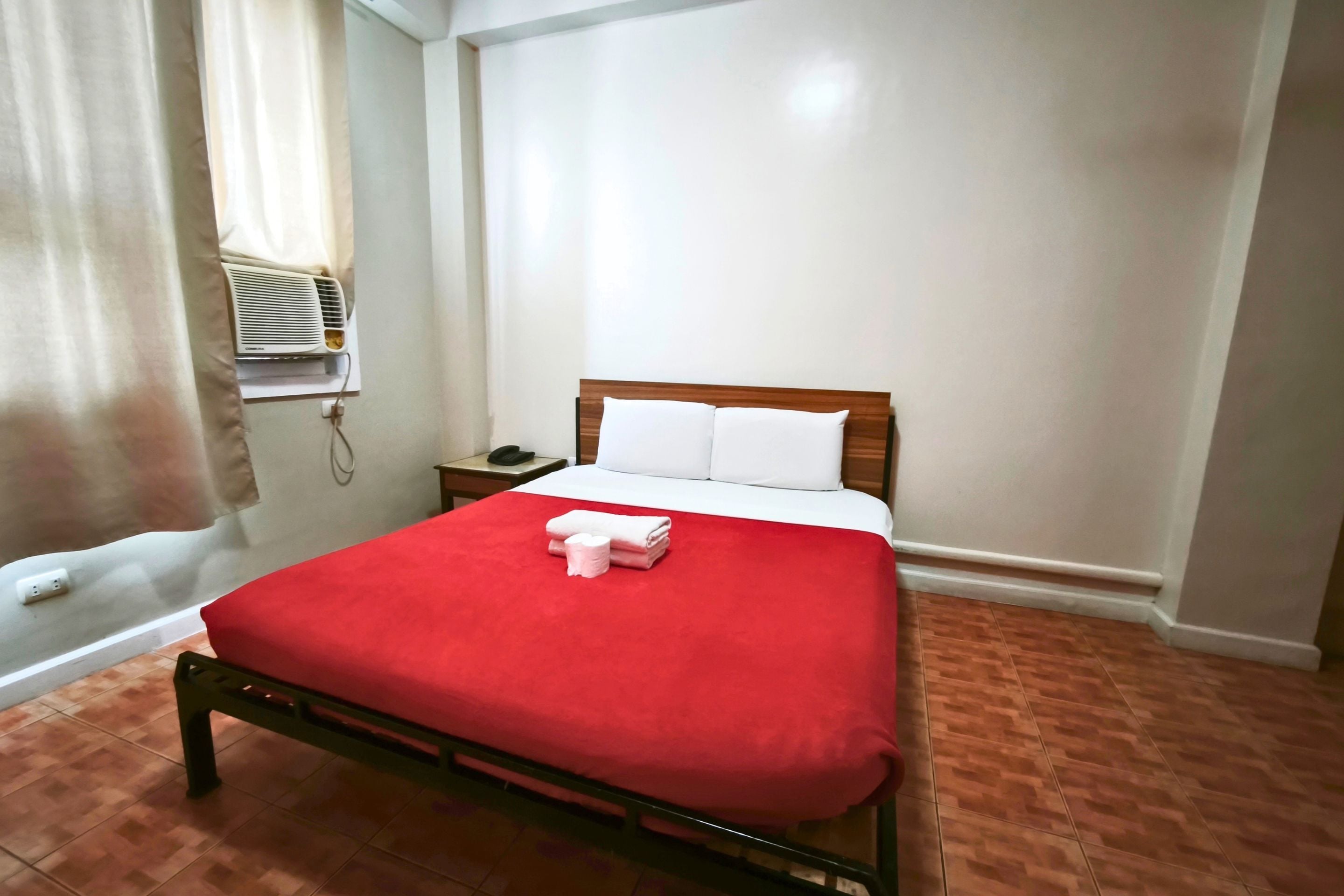 A hotel room with a bed, a bedside table, a telephone, and an air conditioning unit.