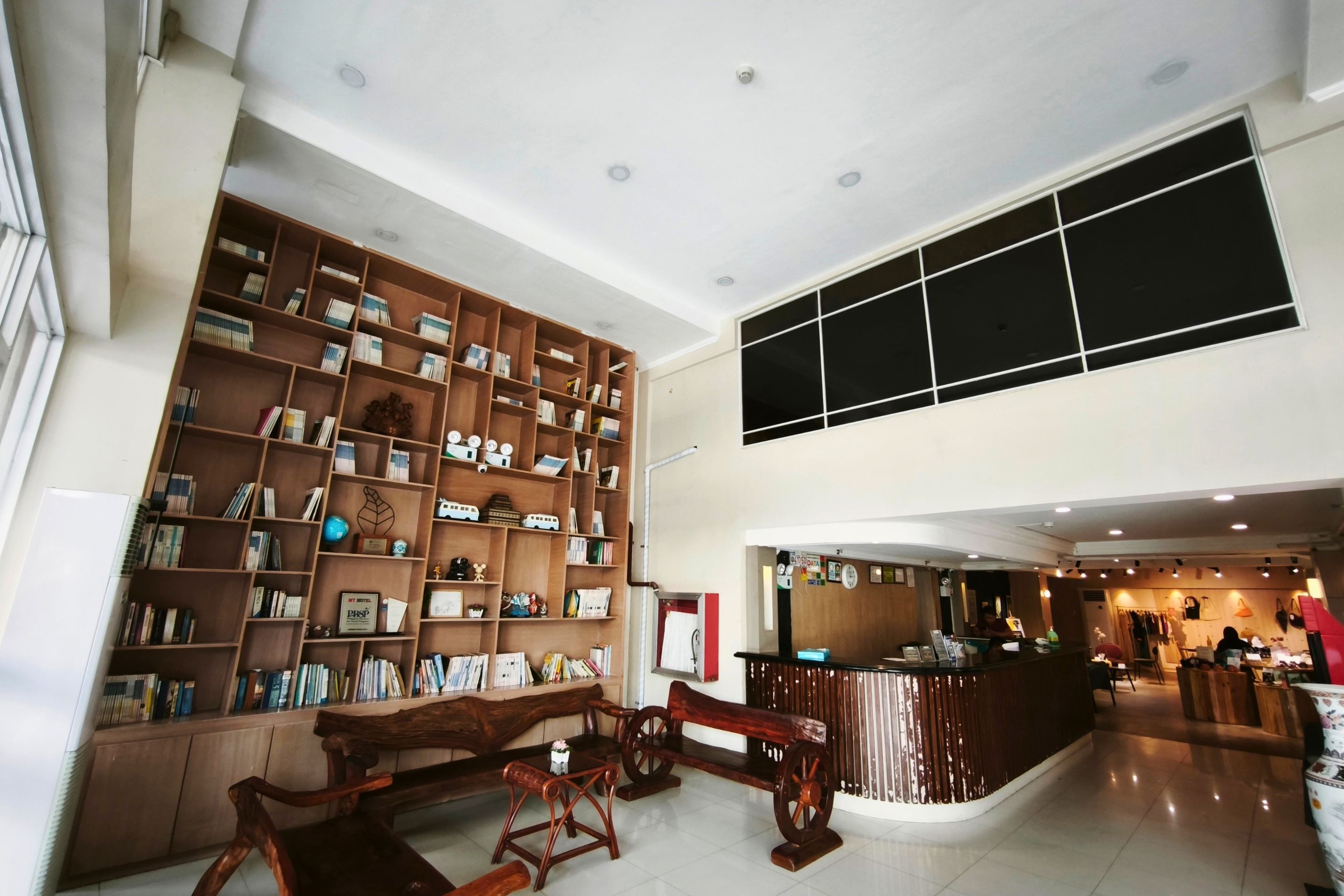 A spacious hotel lobby with a high ceiling, a large wooden bookshelf filled with books and decorative items, a reception desk, and a seating area with a bench and a small table.
