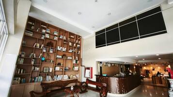 A spacious hotel lobby with a high ceiling, a large wooden bookshelf filled with books and decorative items, a reception desk, and a seating area with a bench and a small table.