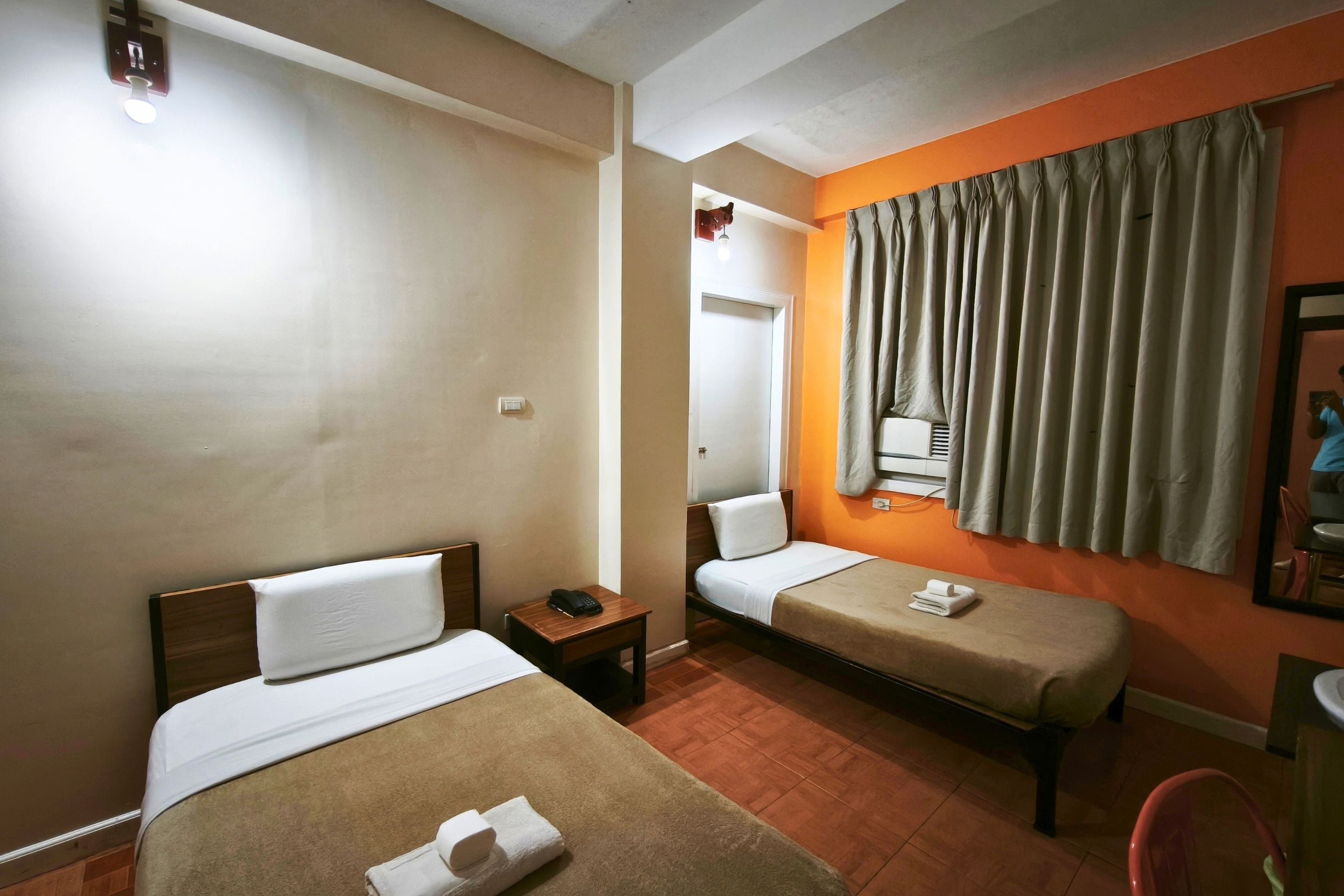 Quarto de hotel com duas camas, uma mesa de cabeceira com telefone, espelho e janela com cortinas.