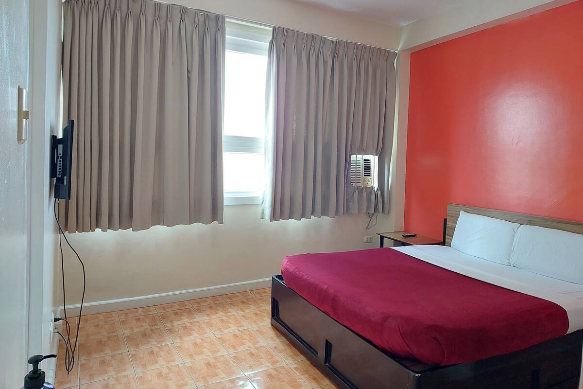 A bedroom with a bed, curtains, a window, an air conditioner, and a red wall.