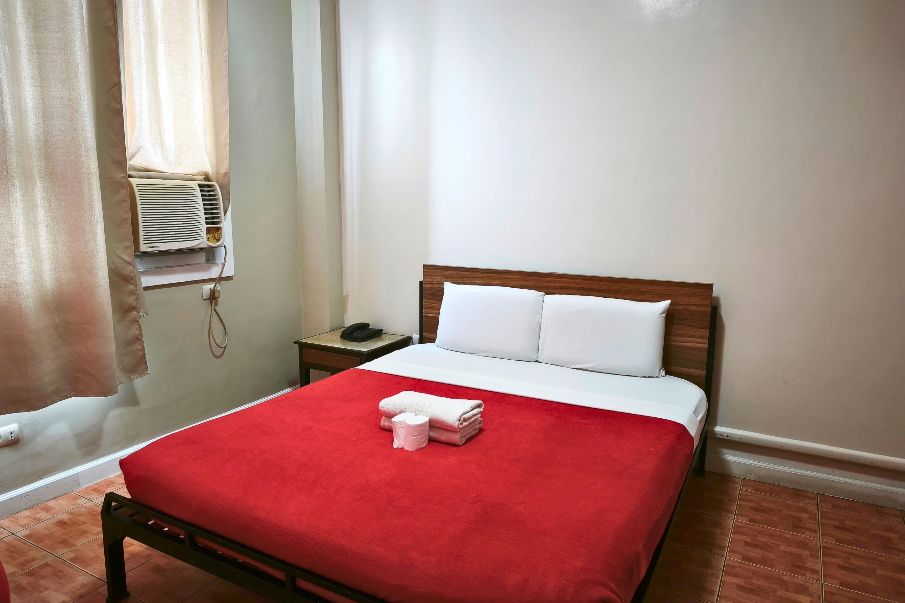 A hotel room with a bed, a bedside table with a phone, and an air conditioning unit.
