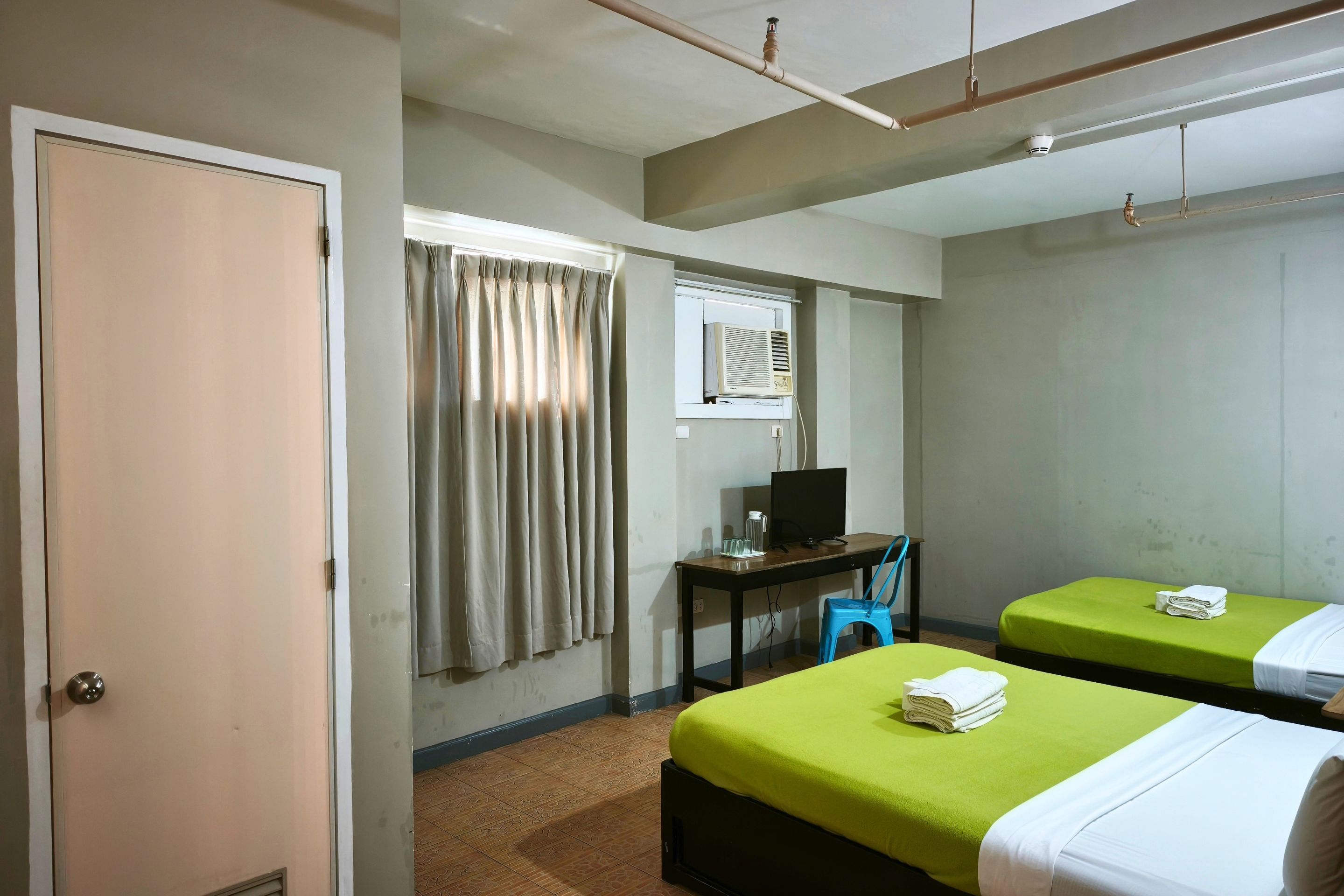 A hotel room with two beds, a desk with a computer, a window with curtains, and a ceiling fan.