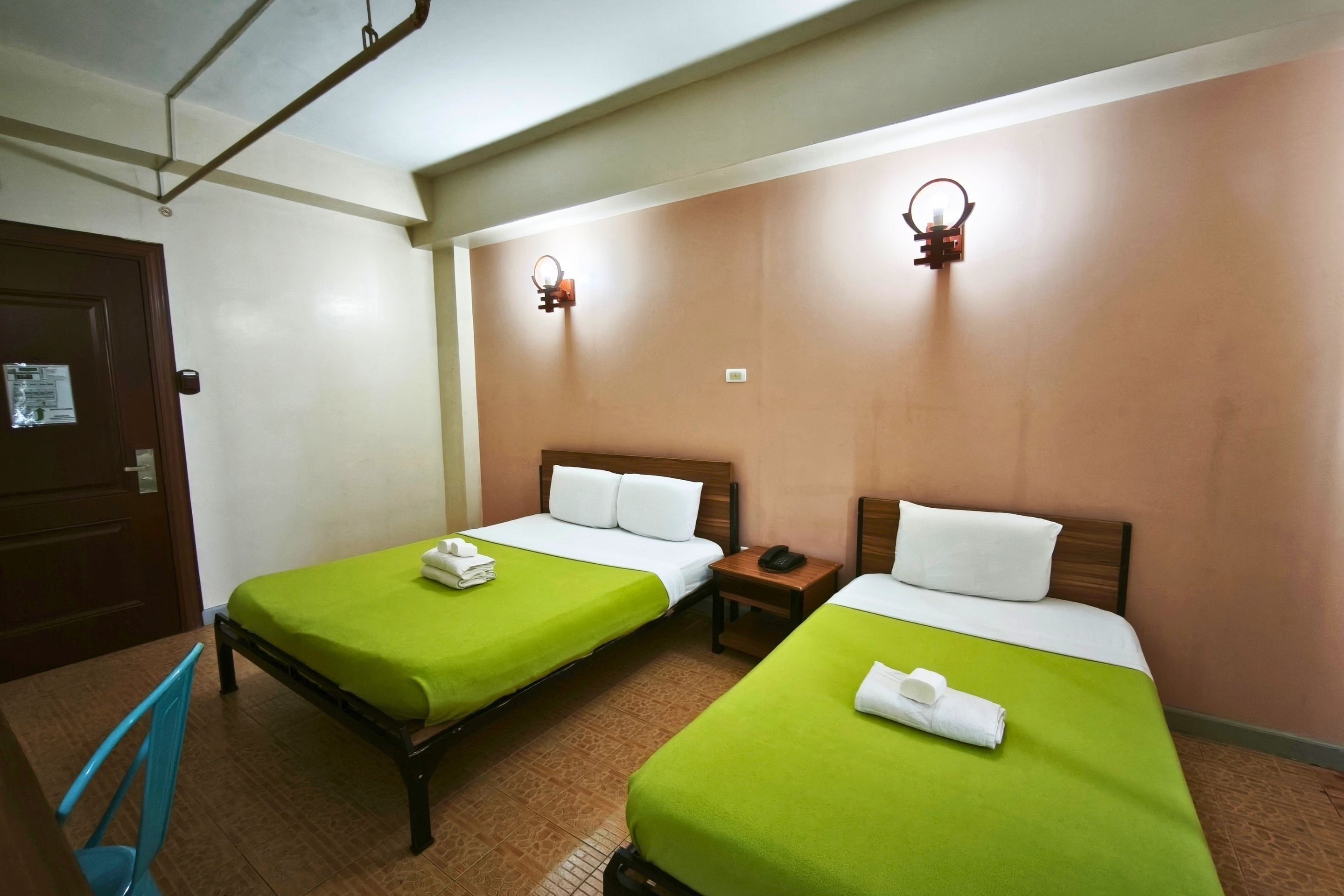 A hotel room with two single beds, each with a green bedspread and white pillows, a wooden nightstand with a phone, and a brown door.