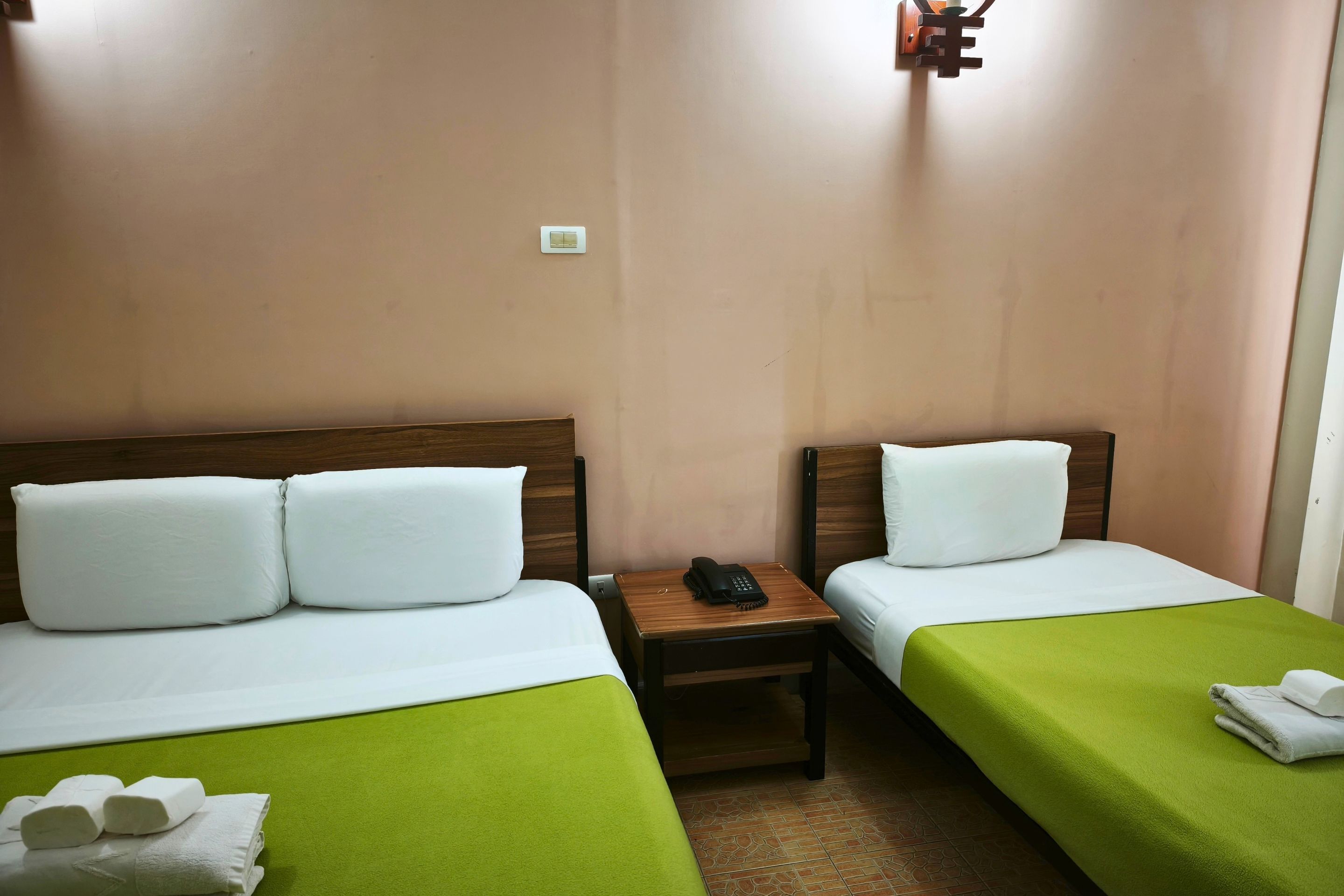 A hotel room with two single beds, a bedside table with a phone, and a wall-mounted light fixture.