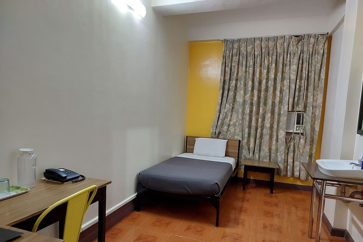 A small, compact room with a bed, a desk, a chair, a sink, and a window with curtains.