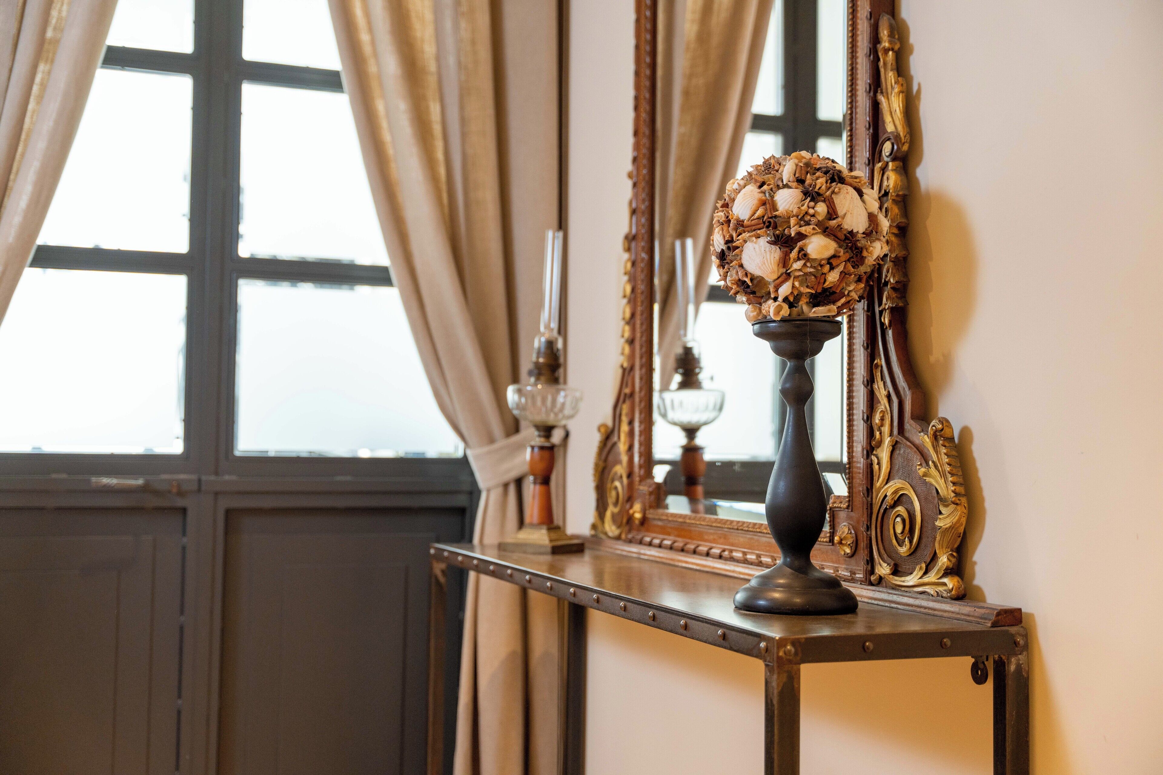 Une pièce au style rétro, dotée d’un grand miroir, d’un vase décoratif contenant des coquillages et d’une fenêtre agrémentée de rideaux.