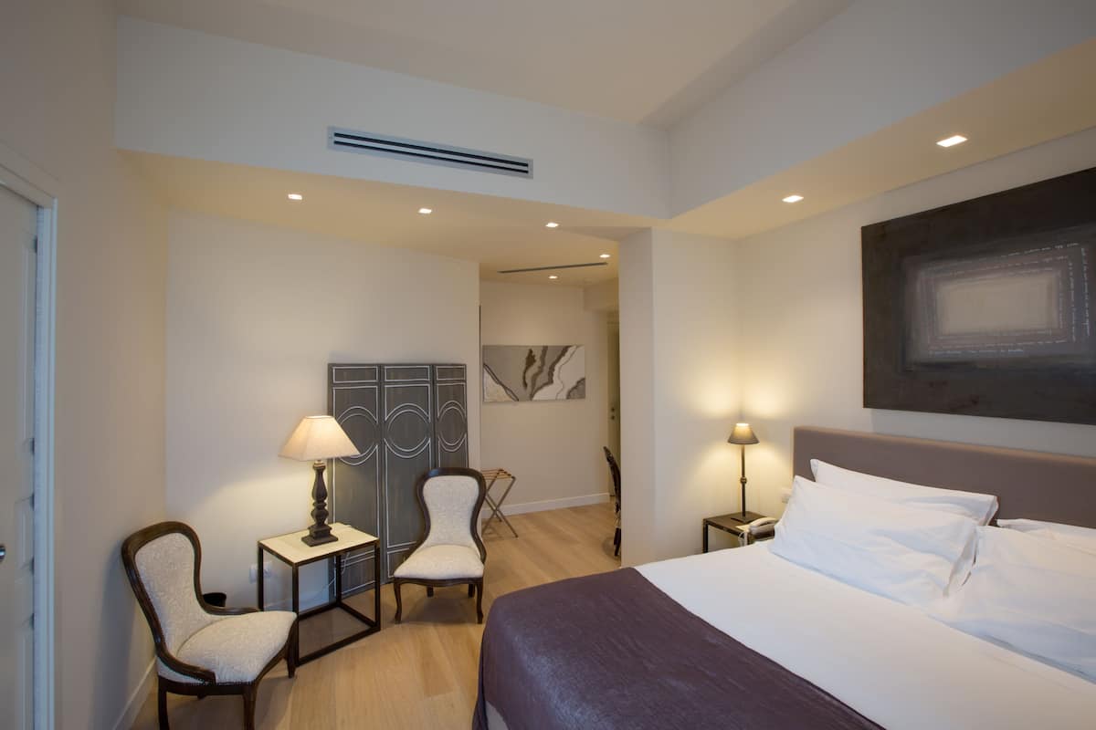 A hotel room with a bed, two chairs, a desk, and a nightstand with a lamp.