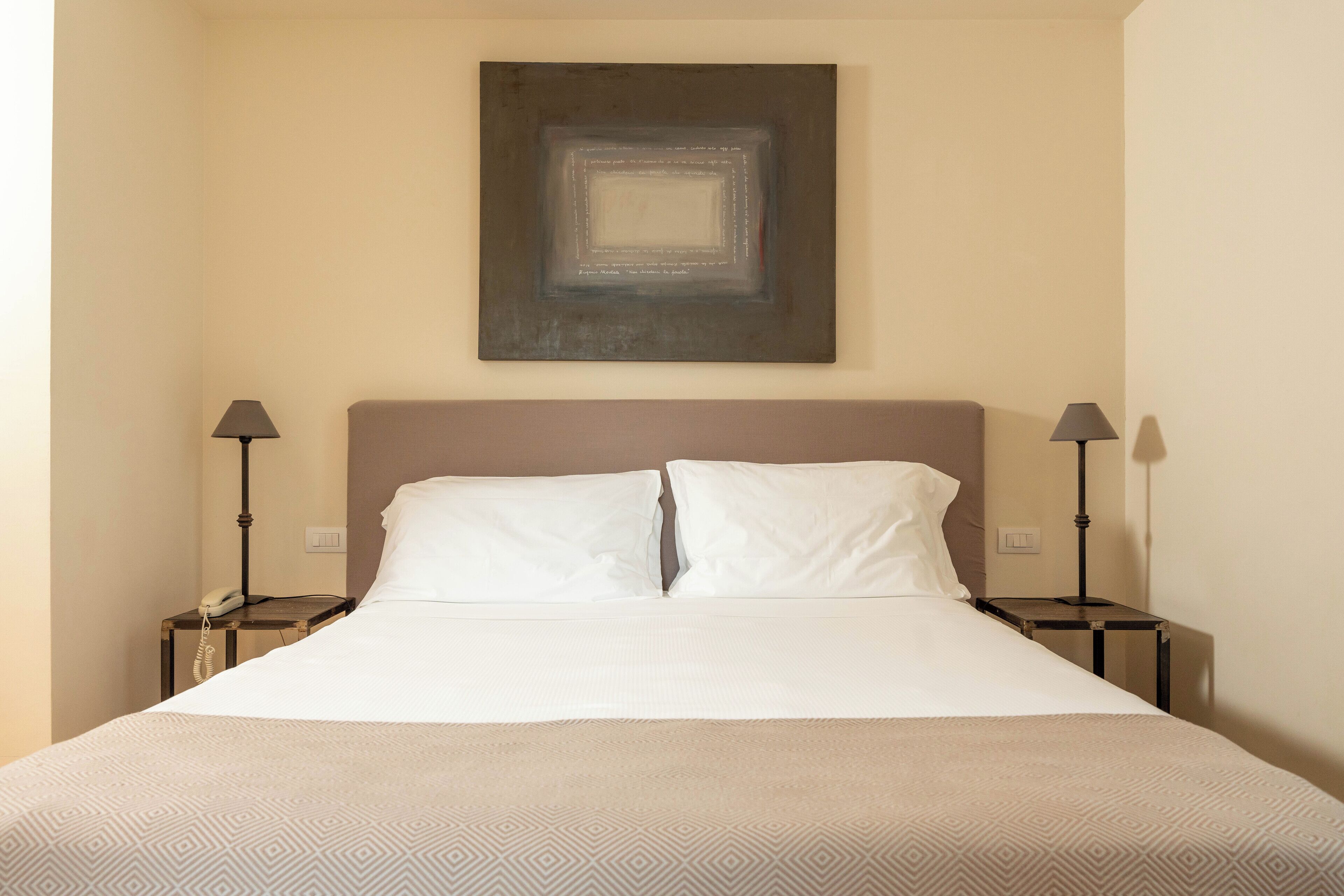 Una camera d'albergo con un letto grande, due comodini con lampade, un'opera d'arte appesa al muro e una parete spoglia.