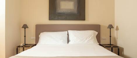 Une chambre d’hôtel avec un grand lit, deux tables de chevet avec des lampes, une œuvre d’art accrochée au mur et un mur sobre.
