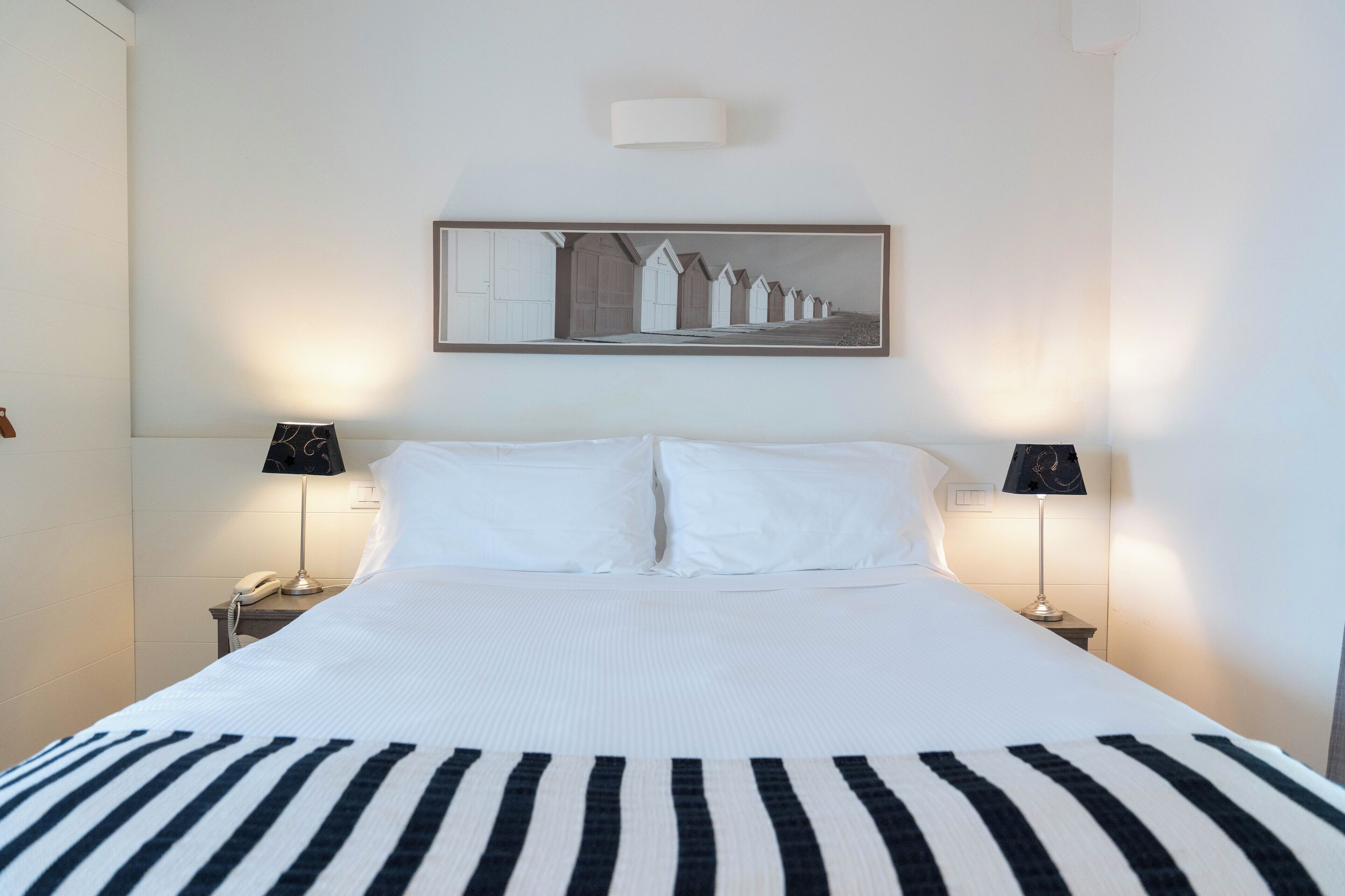 Een net opgemaakt bed met zwart-wit gestreept beddengoed, twee bedlampen, een wandlamp en een ingelijst beeld boven het bed.