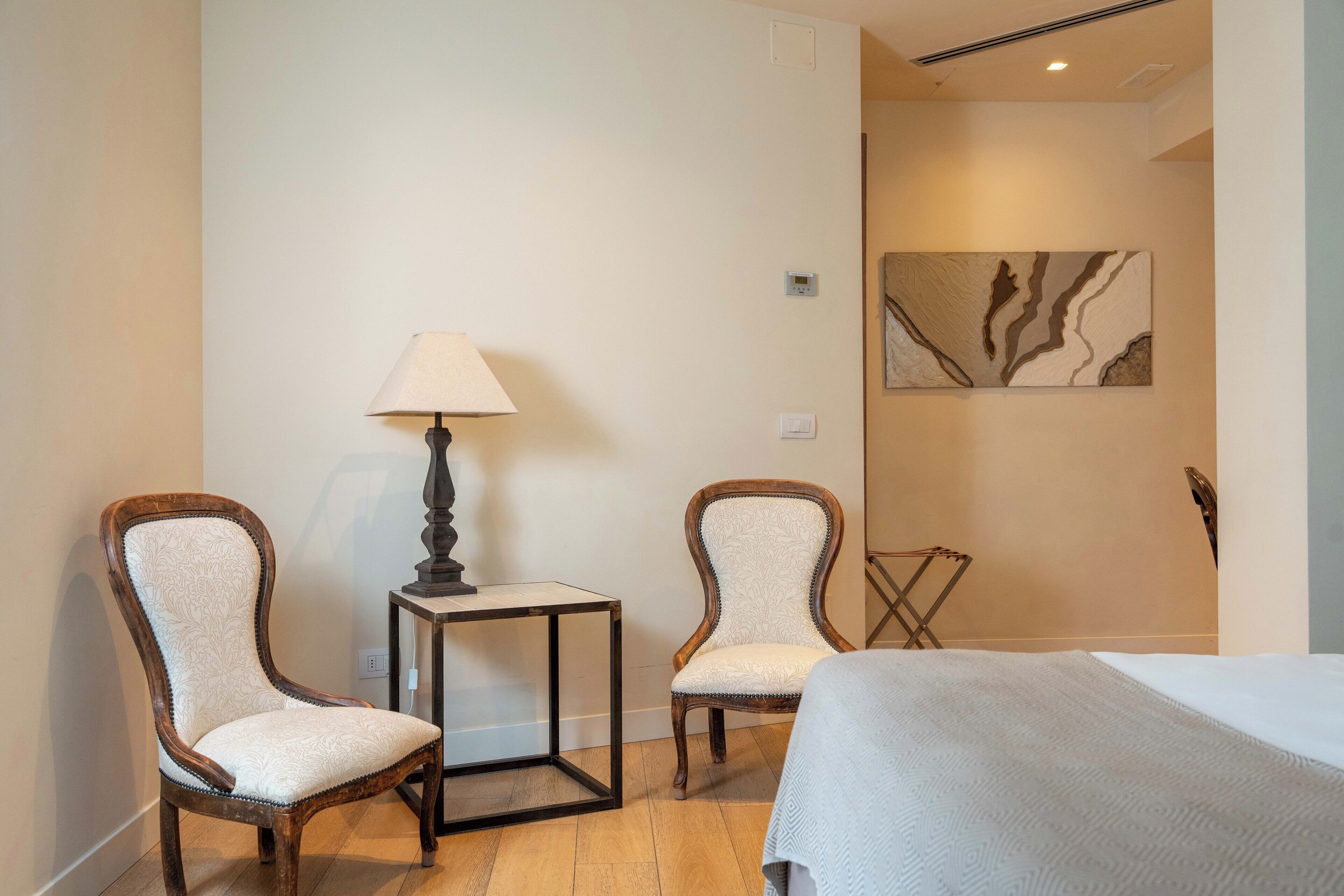 Habitación de hotel con dos sillas de madera, una mesita con lámpara, cama con colcha blanca y una obra de arte colgada en la pared.
