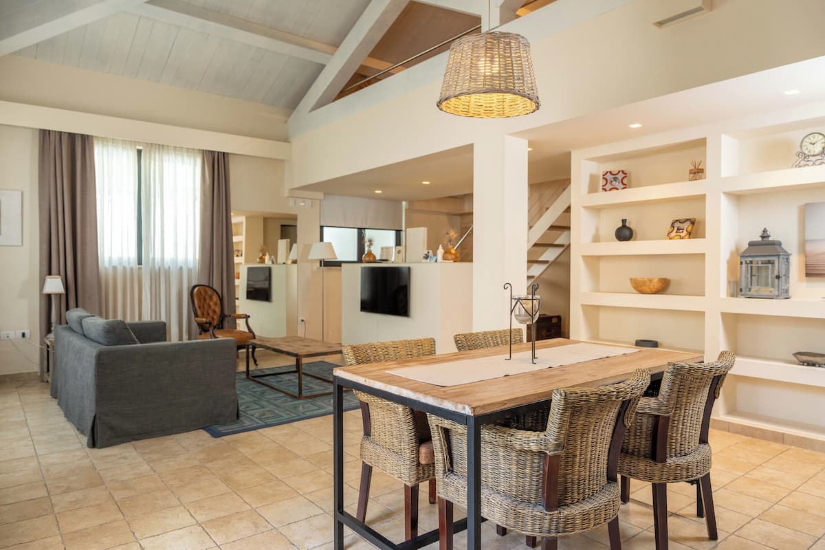 A spacious living area with a dining table, wicker chairs, a sofa, and a staircase leading to another level.