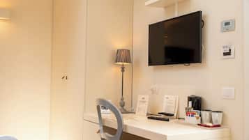 Una camera d'hotel con una scrivania, una sedia, TV a parete, una lampada, telecomando, presa di corrente e termostato fissato al muro.