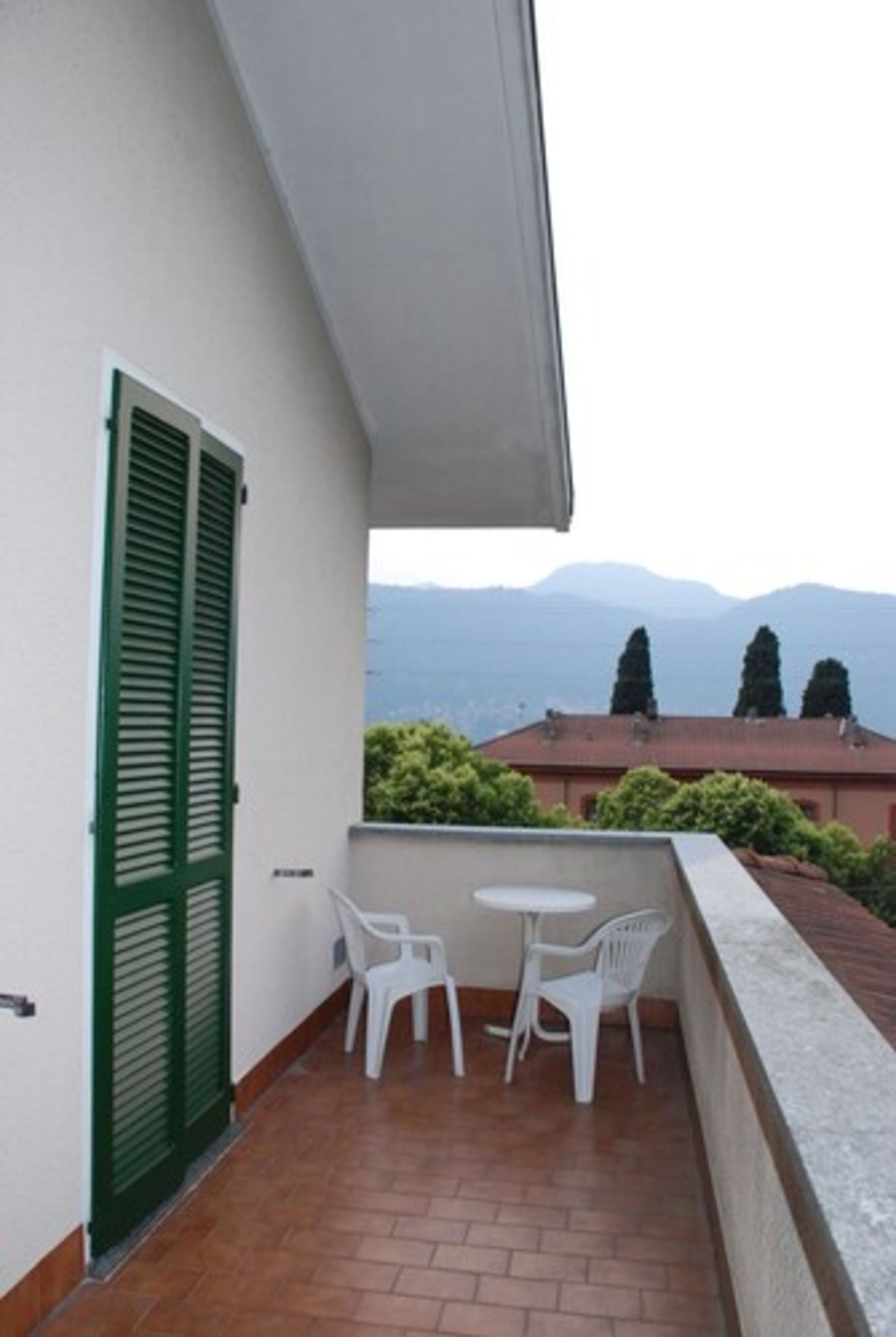 Un balcon avec des chaises en plastique blanches et une petite table, une porte à volets verts et une vue sur des montagnes lointaines.