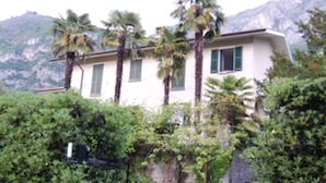 A house with a stone pathway leading to a gate, surrounded by greenery and palm trees, with mountains in the background. - Bed and Breakfast Casa Nini (Lierna)