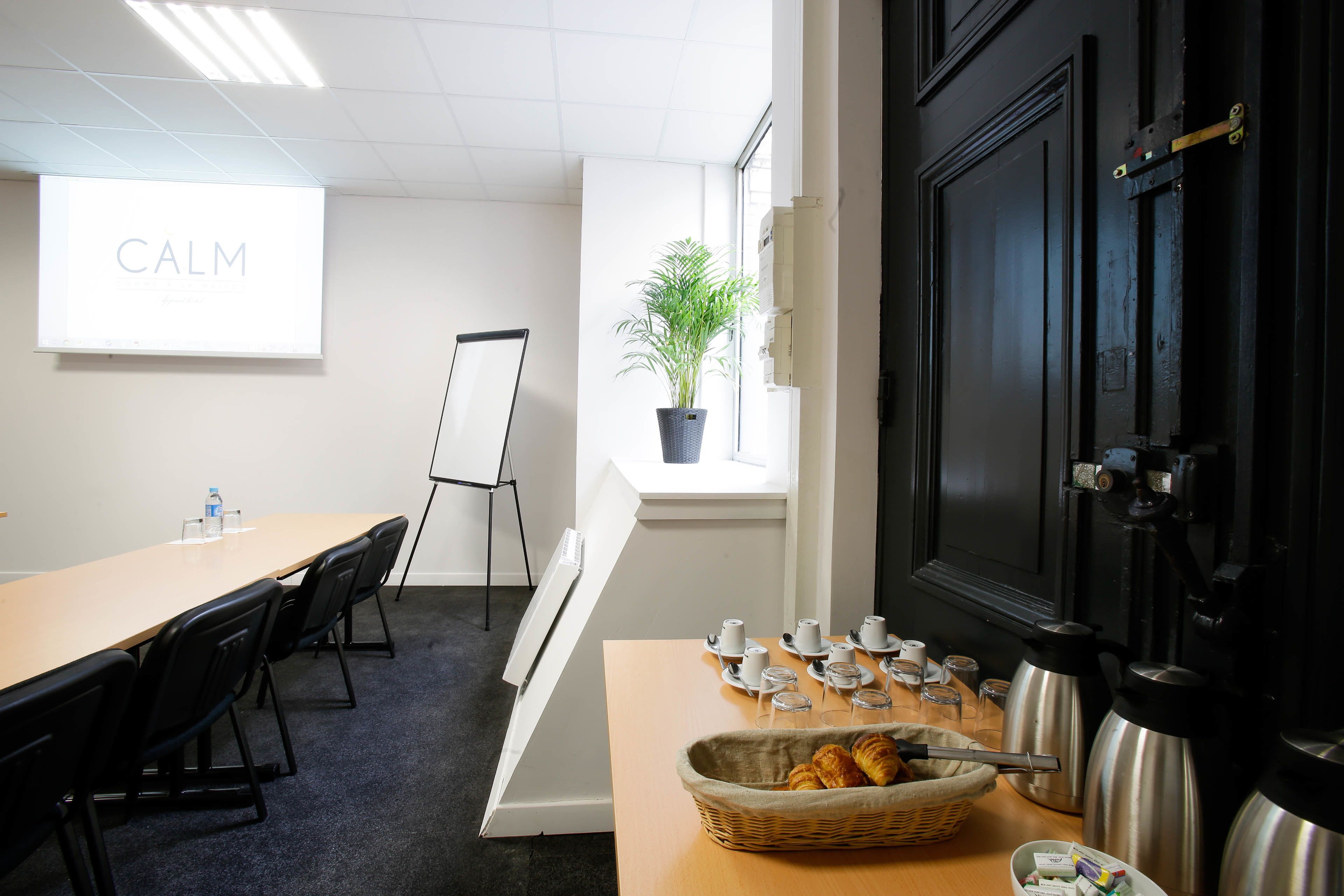 Une salle de réunion avec une longue table, des chaises, un écran de projection affichant le mot « CALME », un tableau blanc, une fenêtre avec une plante, et une table avec des tasses à thé, des soucoupes et un panier de croissants.