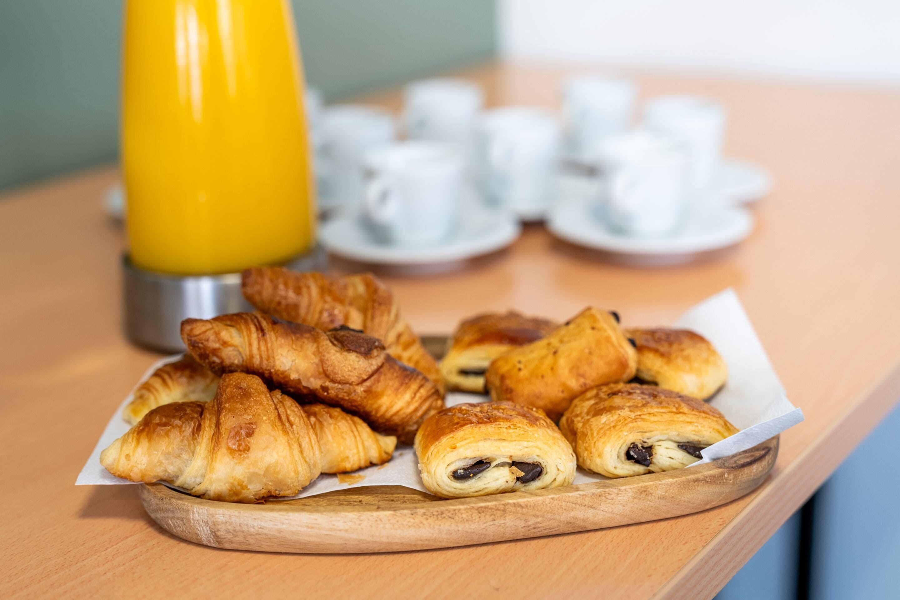Een houten dienblad met croissants en een gele beker, waarschijnlijk voor sap, op een tafel. Op de achtergrond witte kopjes en schotels.