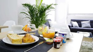 A breakfast setup with toast, croissants, fruit, juice, and yogurt on a table in a room with a plant and a sofa. - CALM Appart' & Hostel (Lille)