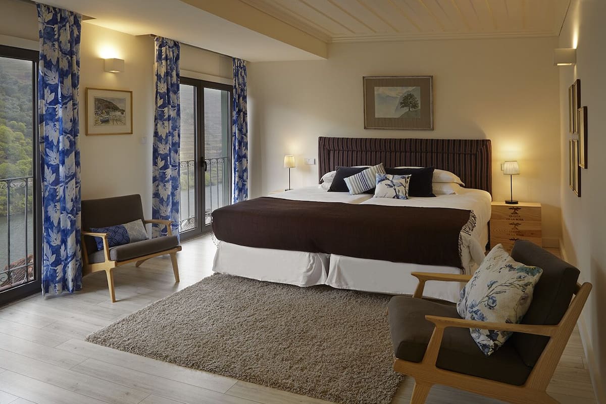 A bedroom with a large bed, two bedside tables with lamps, a chair, a view of the outdoors through a window with curtains, and a framed picture on the wall.
