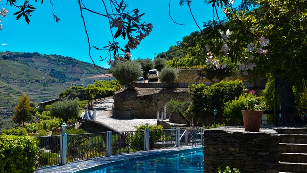 A pool area with a stone wall, a white fence, and a view of a hillside with a building and a car parked on a slope.