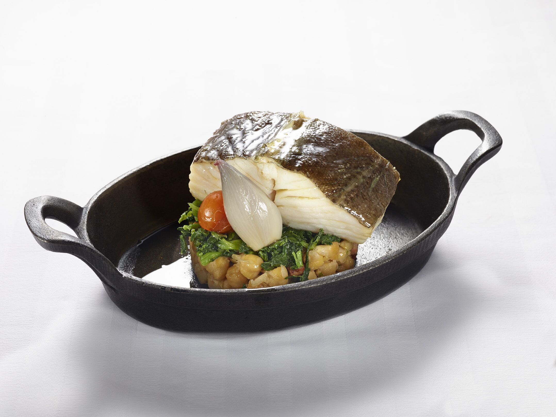 Une poêle en fonte contenant un morceau de poisson, des légumes et une tomate, le tout posé sur une surface blanche.