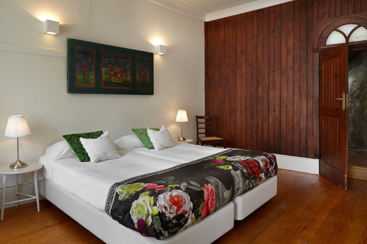 A bedroom with a wooden floor, a bed with white bedding and green pillows, two bedside lamps, and a wooden door.