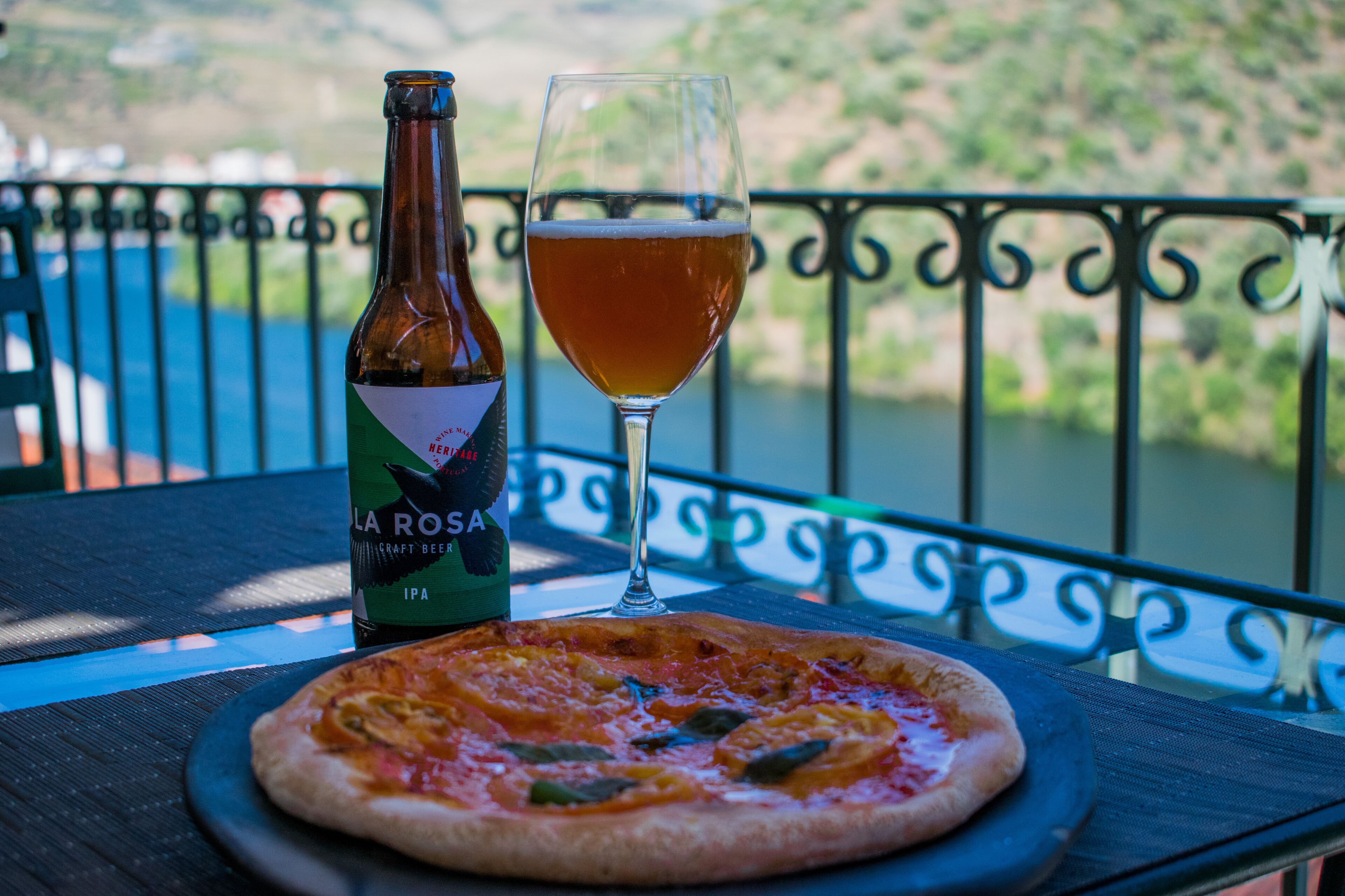 A bottle of La Rosa IPA beer, a glass of beer, and a pizza on a table with a view of a river and hills.